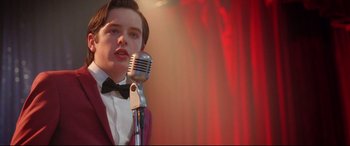 Movie still from “Sing Street” (2016), directed by John Carney – A young man in a suit and bow tie talking into a microphone; Close Up shot, Low angle