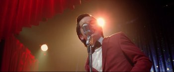 Movie still from “Sing Street” (2016), directed by John Carney – A man in a red jacket is singing into a microphone; Medium shot, Low angle