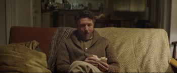 Movie still from “Sing Street” (2016), directed by John Carney – A man sitting in a chair eating a sandwich; Medium shot, High angle