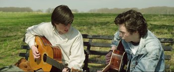 Movie still from “Sing Street” (2016), directed by John Carney – Two young men sitting on a park bench with guitars; Medium shot, Over the shoulder angle