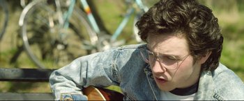 Movie still from “Sing Street” (2016), directed by John Carney – A man with glasses is leaning on a guitar; Close Up shot, High angle