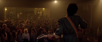 Movie still from “Sing Street” (2016), directed by John Carney – A man holding a guitar in front of an audience; Wide shot, Over the shoulder angle