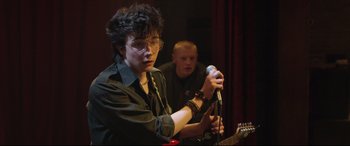 Movie still from “Sing Street” (2016), directed by John Carney – A man holding a microphone while standing next to another man; Medium shot, Over the shoulder angle