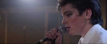 Movie still from “Sing Street” (2016), directed by John Carney – A person holding a microphone up to their mouth; Close Up shot, Over the shoulder angle