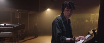 Movie still from “Sing Street” (2016), directed by John Carney – A young man standing in a dark room; Medium shot, Over the shoulder angle