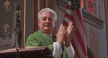 Movie still from “Sister Act” (1992), directed by Emile Ardolino – An older man wearing a green robe clapping in front of an american flag; Medium shot, Low angle