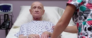 Movie still from “Sisters” (2015), directed by Jason Moore – An older man in a hospital bed with a nurse touching his arm; Close Up shot, Over the shoulder angle