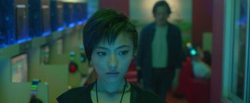 A woman with short black hair is looking at the camera while a man is standing in the background