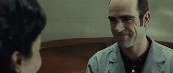 Movie still from “Sleep Tight” (2011), directed by Jaume Balagueró – A man with a bald head smiling for the camera; Close Up shot, Over the shoulder angle