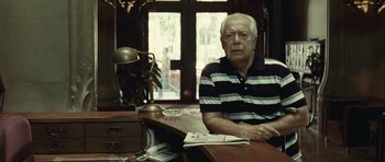 Movie still from “Sleep Tight” (2011), directed by Jaume Balagueró – An older man sitting at a desk in front of a window; Medium shot, Low angle