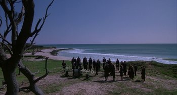 Movie still from “Sleeping with the Enemy” (1991), directed by Joseph Ruben – A group of people standing on top of a grass covered beach; Extreme Wide shot, High angle