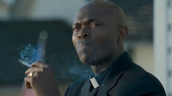 Movie still from “Slither” (2006), directed by James Gunn – A man in a priest's outfit smoking a cigarette; Close Up shot, Low angle