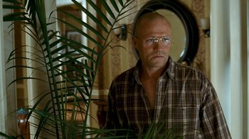 Movie still from “Slither” (2006), directed by James Gunn – A bald man wearing glasses and a plaid shirt is standing in front of a plant; Medium shot, Low angle