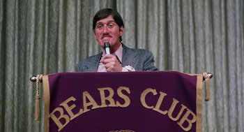 Movie still from “Smile” (1975), directed by Michael Ritchie – A man is speaking at a podium at a bears club event; Close Up shot, Low angle
