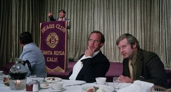 Movie still from “Smile” (1975), directed by Michael Ritchie – Two men sitting at a table with a banner in the background; Medium shot, Low angle