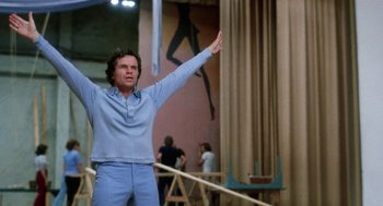 Movie still from “Smile” (1975), directed by Michael Ritchie – A man standing in front of a wall holding his arms up; Medium shot, Low angle