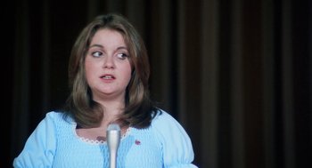 Movie still from “Smile” (1975), directed by Michael Ritchie – A woman in a blue dress is speaking into a microphone; Close Up shot, Over the shoulder angle