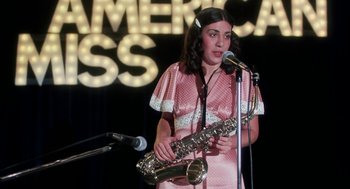 Movie still from “Smile” (1975), directed by Michael Ritchie – A woman holding a saxophone in front of a microphone; Medium shot, High angle