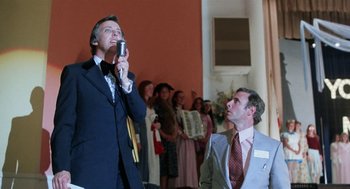 Movie still from “Smile” (1975), directed by Michael Ritchie – A man in a suit and tie holding up a microphone; Medium shot, Low angle