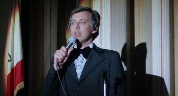 Movie still from “Smile” (1975), directed by Michael Ritchie – A man in a tuxedo holding a microphone; Close Up shot, Low angle