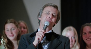 Movie still from “Smile” (1975), directed by Michael Ritchie – A man in a black suit is holding a microphone; Close Up shot, Low angle