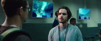 Movie still from “Snowden” (2016), directed by Oliver Stone – A man with long brown hair standing in front of a television; Close Up shot, Over the shoulder angle