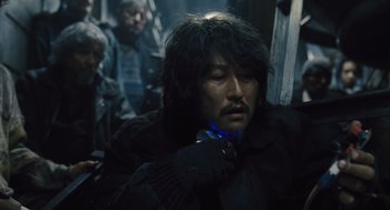 Movie still from “Snowpiercer” (2013), directed by Bong Joon Ho – A man with long black hair and a beard is sitting down; Close Up shot, Over the shoulder angle