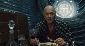 Movie still from “Snowpiercer” (2013), directed by Bong Joon Ho – An older man sitting at a table with a plate of food and a glass of red wine; Close Up shot, Over the shoulder angle