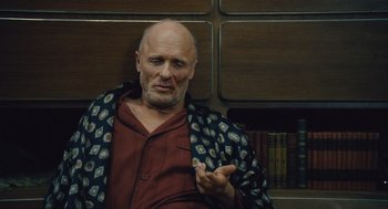 Movie still from “Snowpiercer” (2013), directed by Bong Joon Ho – An older man is sitting on a chair in front of a book case; Close Up shot, Over the shoulder angle