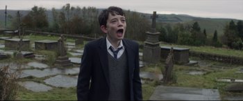 Movie still from “A Monster Calls” (2016), directed by J.A. Bayona – A young boy in a suit and tie standing next to a grave; Medium shot, Over the shoulder angle
