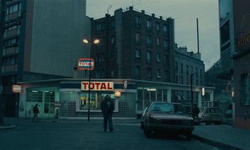 Movie still from “So Long, Stooge” (1983), directed by Claude Berri – A man standing on the side of the road near a gas station; Extreme Wide shot, High angle