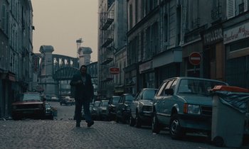 Movie still from “So Long, Stooge” (1983), directed by Claude Berri – A man walking down a street next to parked cars; Wide shot, High angle