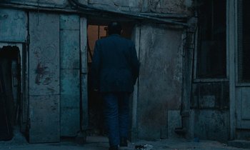 Movie still from “So Long, Stooge” (1983), directed by Claude Berri – A man in a suit and hat walking in front of an open door; Wide shot, Over the shoulder angle