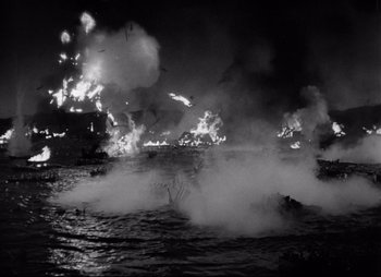 Movie still from “So Proudly We Hail” (1990), directed by Lionel Chetwynd – A black - and - white photo of a boat on fire; Extreme Wide shot, High angle