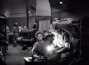 Movie still from “So Proudly We Hail” (1990), directed by Lionel Chetwynd – Two men are sitting in a room with a lamp; Wide shot, High angle