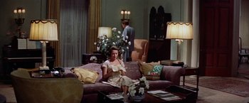 Movie still from “Some Came Running” (1958), directed by Vincente Minnelli – A woman sitting on top of a purple couch in a living room; Wide shot, High angle