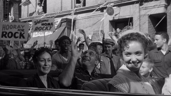 Movie still from “Somebody Up There Likes Me” (1956), directed by Robert Wise – A black and white photo of people in a parade; Medium shot, Low angle