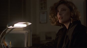 Movie still from “Someone to Watch Over Me” (1987), directed by Ridley Scott – A woman sitting at a table in a dark room; Close Up shot, Over the shoulder angle