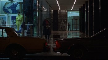Movie still from “Someone to Watch Over Me” (1987), directed by Ridley Scott – A person standing in a building with a car in front of it; Extreme Wide shot, Over the shoulder angle