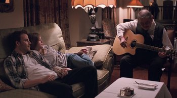 Movie still from “Somewhere” (2010), directed by Sofia Coppola – Two people sitting on a couch with a man playing a guitar; Medium shot, Over the shoulder angle