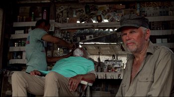 Movie still from “Sorcerer” (1977), directed by William Friedkin – A man sitting in a barber shop with a man cutting his hair; Medium shot, Over the shoulder angle