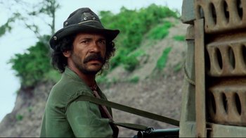 Movie still from “Sorcerer” (1977), directed by William Friedkin – A man with a hat and a beard is leaning against a pole; Close Up shot, Low angle