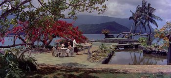 Movie still from “South Pacific” (1958), directed by Joshua Logan – A group of people sitting at a table near a body of water; Extreme Wide shot, High angle