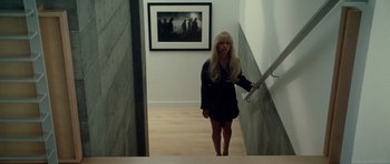 Movie still from “Southland Tales” (2006), directed by Richard Kelly – A woman standing on the bottom of a flight of stairs in front of a picture on the wall; Wide shot, High angle