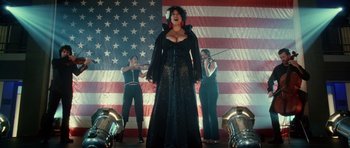 Movie still from “Southland Tales” (2006), directed by Richard Kelly – A woman in a black dress singing in front of an american flag; Wide shot, Low angle