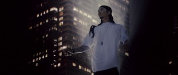 Movie still from “Southland Tales” (2006), directed by Richard Kelly – A man in a white shirt and a black hat is standing in front of a tall building; Medium shot, Low angle