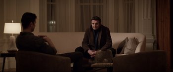 Movie still from “A Walk Among the Tombstones” (2014), directed by Scott Frank – A man sitting on top of a couch in a living room; Medium shot, Over the shoulder angle