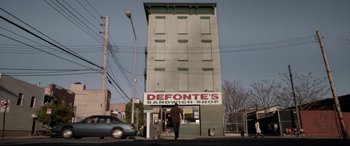 Movie still from “A Walk Among the Tombstones” (2014), directed by Scott Frank – A man walking down the street in front of a sandwich shop; Wide shot, Low angle