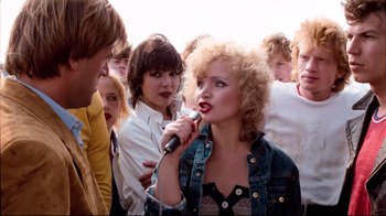 Movie still from “Spetters” (1980), directed by Paul Verhoeven – A group of people standing around a woman with a microphone; Medium shot, Over the shoulder angle