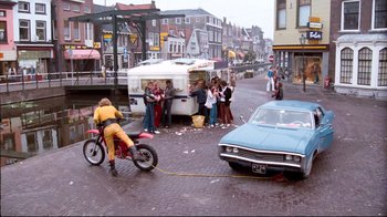Movie still from “Spetters” (1980), directed by Paul Verhoeven – A group of people standing next to a car and a motorcycle; Wide shot, High angle